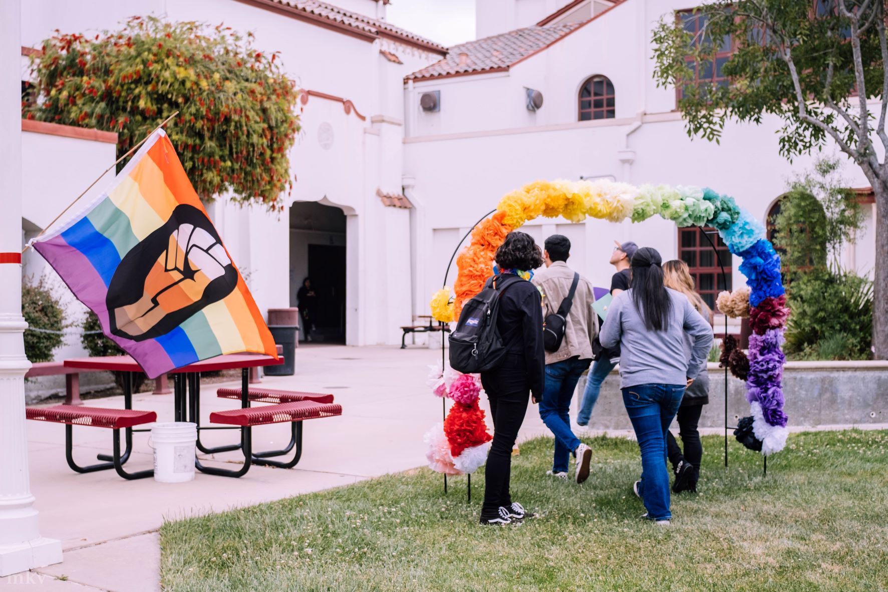 people walk under a rainbow balloon arch near an LGBTQ+ Pride flag