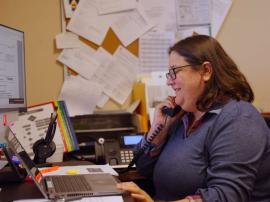 a person talks on a phone at a desk with a computer and a bulletin board