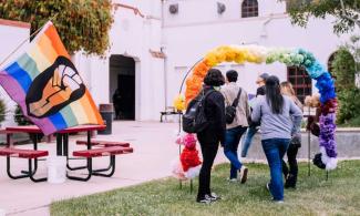 people walk under a rainbow balloon arch near an LGBTQ+ Pride flag