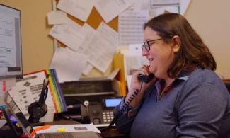 a person talks on a phone at a desk with a computer and a bulletin board