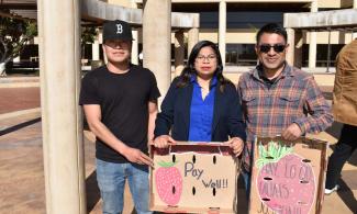 three people stand in front of concrete columns holding signs about fair wages